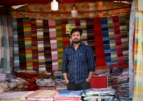 Take your business global - we take care of tech Man standing in colorful textile market stall featuring various fabric patterns.